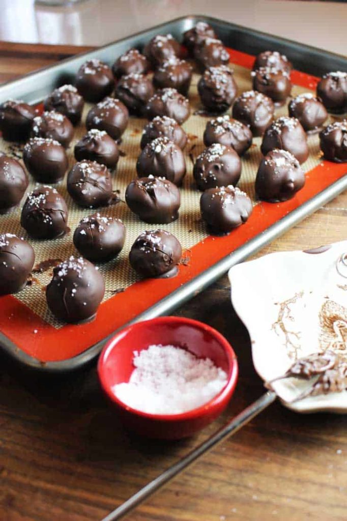 Dark chocolate truffles with caramel, just dipped in melted tempered chocolate and sprinkled with Fleur de sel, on a sheet pan lined with silpat