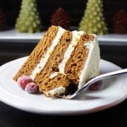 Slice of gingerbread cake with brown butter cream cheese frosting on a plate, decorated with sugared cranberries