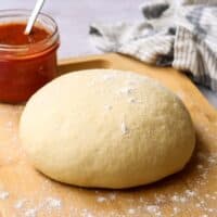 A ball of raw pizza dough on a wooden cutting board with a jar of pizza sauce in the background.