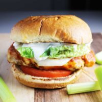 A Buffalo chicken burger with cheese, ranch, tomato and lettuce on a wooden cutting board