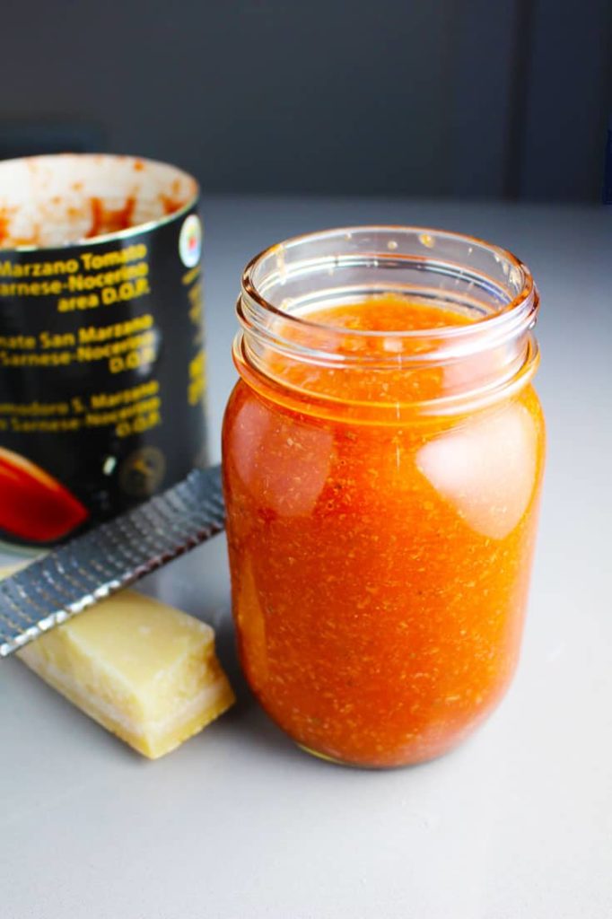 Pizza sauce in a mason jar with parmesan cheese, a microplane grater, and a can of tomatoes in the background