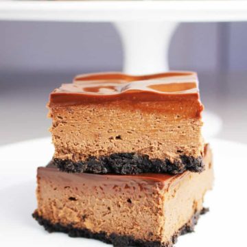 Two triple chocolate cheesecake bars stacked on top of one another on a white plate with more bars on a cake stand in the background