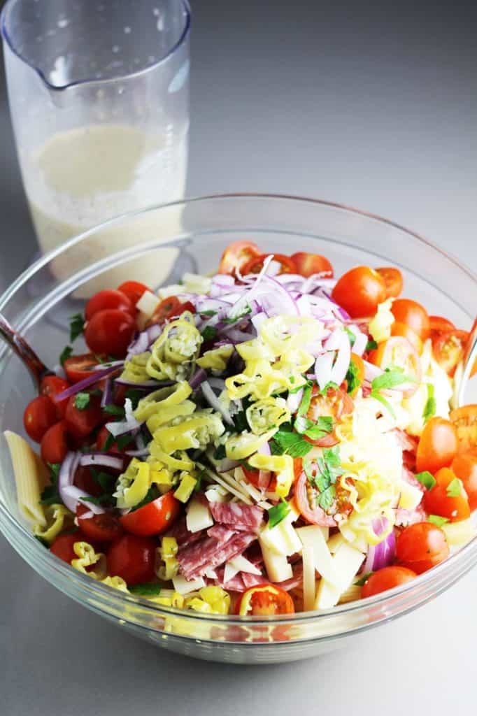 Ingredients for Italian pasta salad in a large salad bowl with prepared dressing in the background