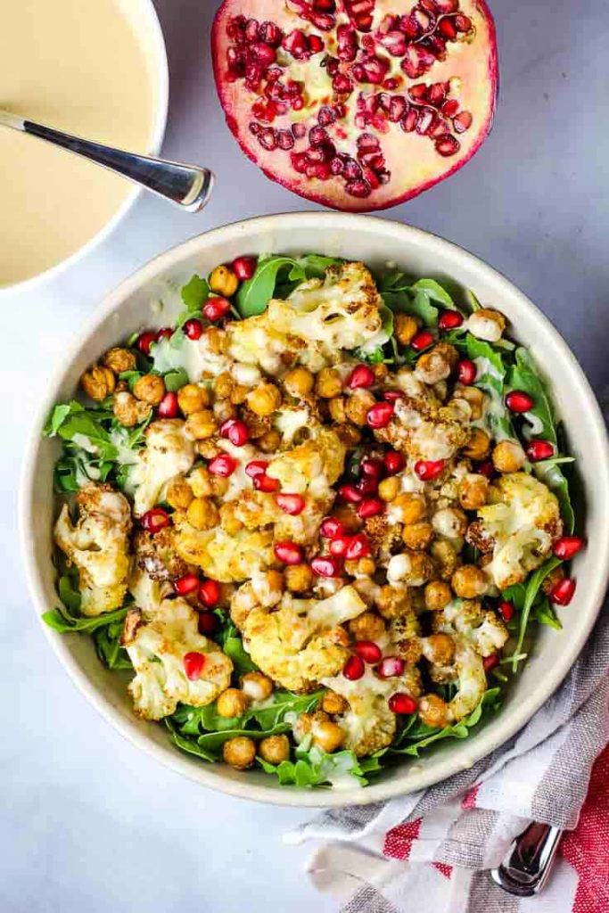 A bowl of roasted cauliflower salad with tahini dressing and a pomegranate half in background