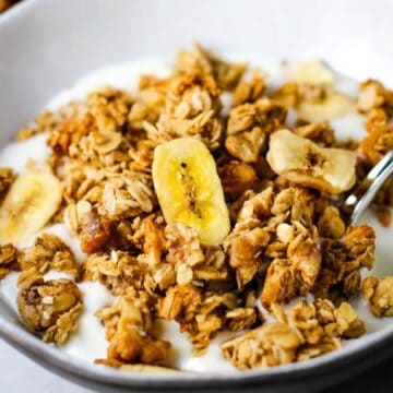 Closeup of a bowl of granola with banana chips over vanilla yogurt with a spoon digging in.