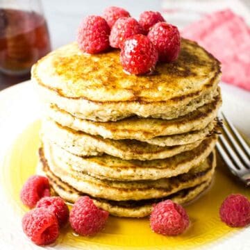 A stack of oat flour pancakes garnished with raspberries and syrup.
