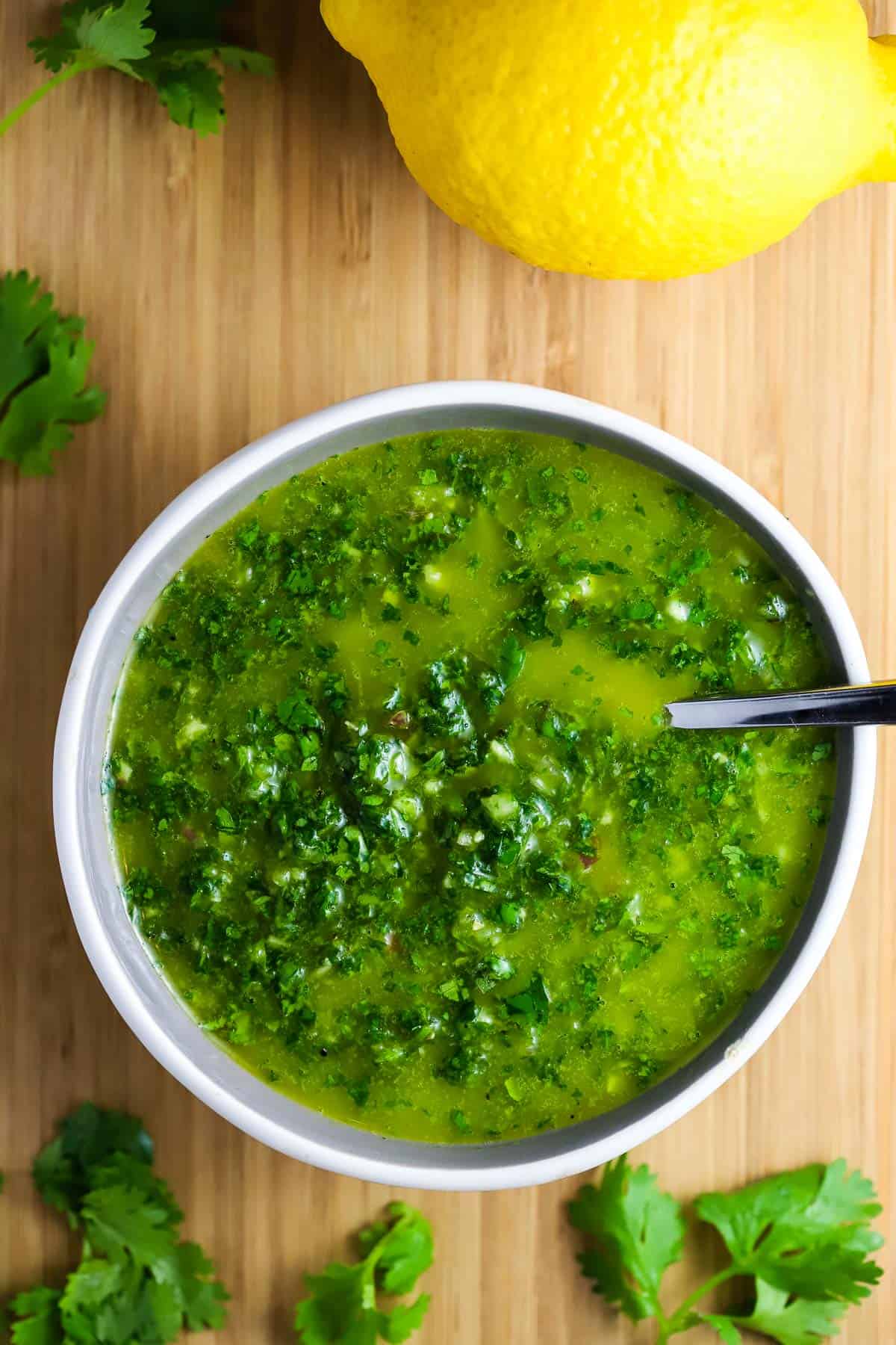 Overhead view of a bowl of chimichurri with a spoon in it and a whole lemon in the background.