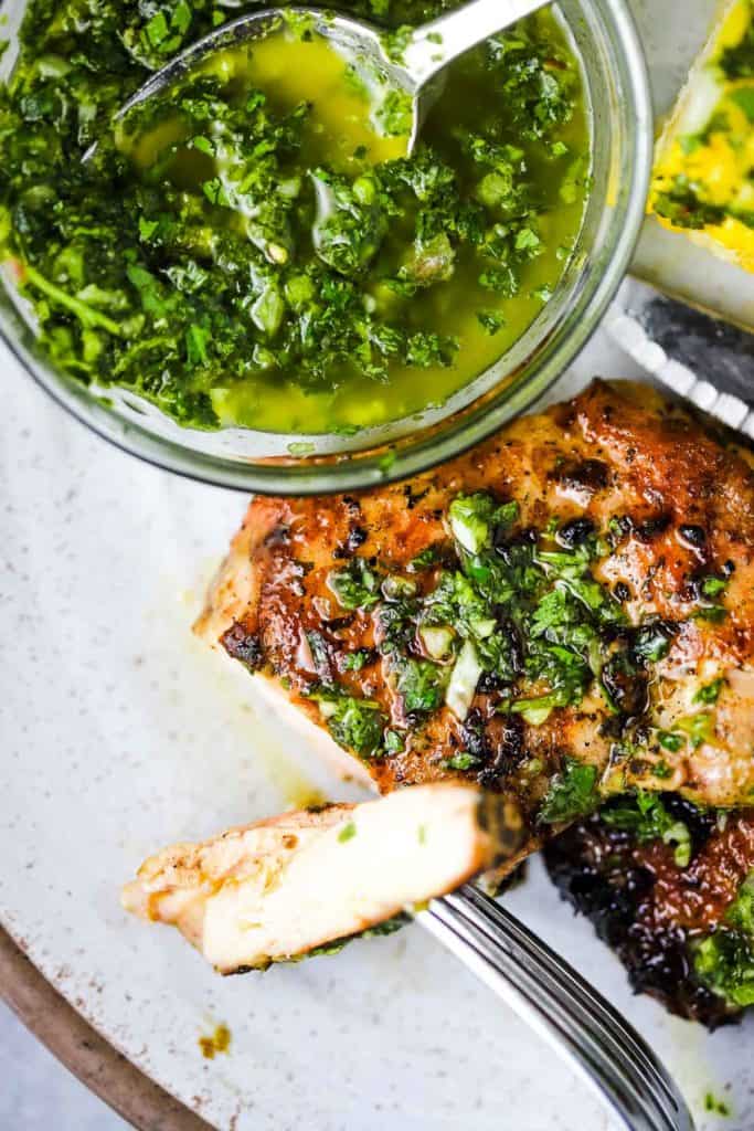 A chicken thigh with a slice on a fork, and a bowl of chimichurri