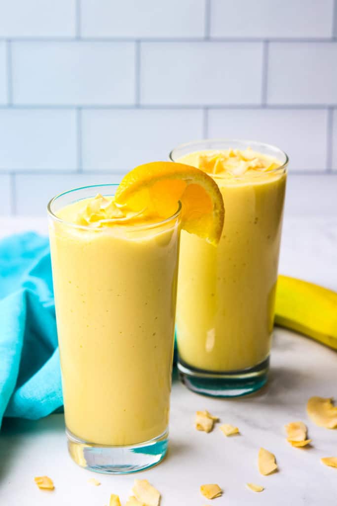 Two mango pineapple smoothies in tall glasses garnished with coconut chips and orange slices