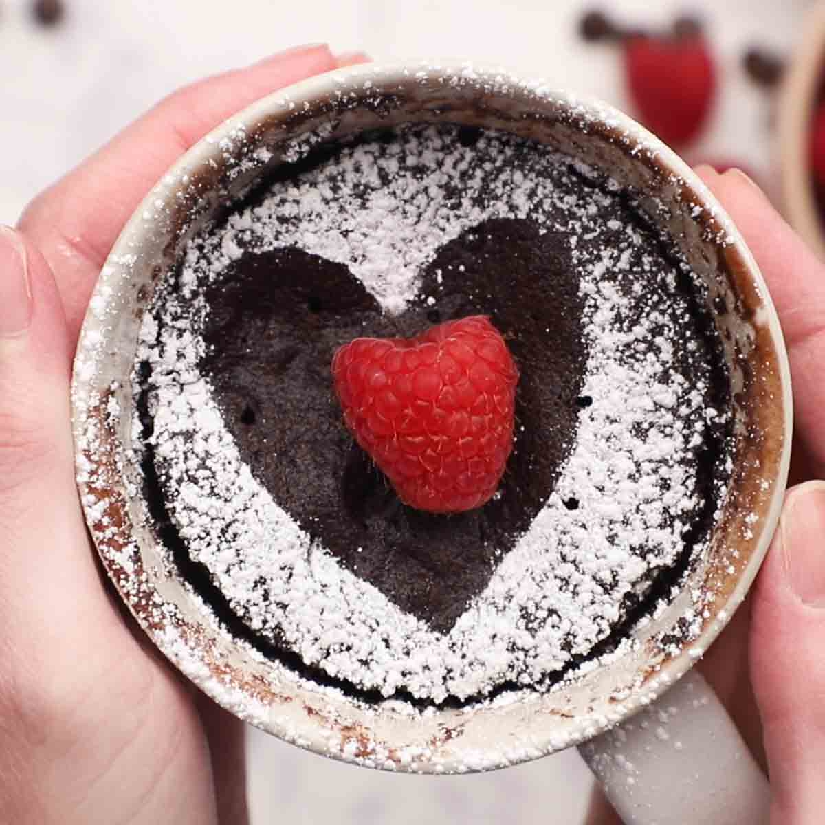 Two hands holding a chocolate mug cake decorated with a powdered sugar hear and raspberry
