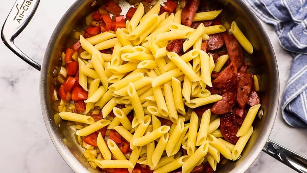 Penne pasta, sausages, and vegetables in a frying pan.