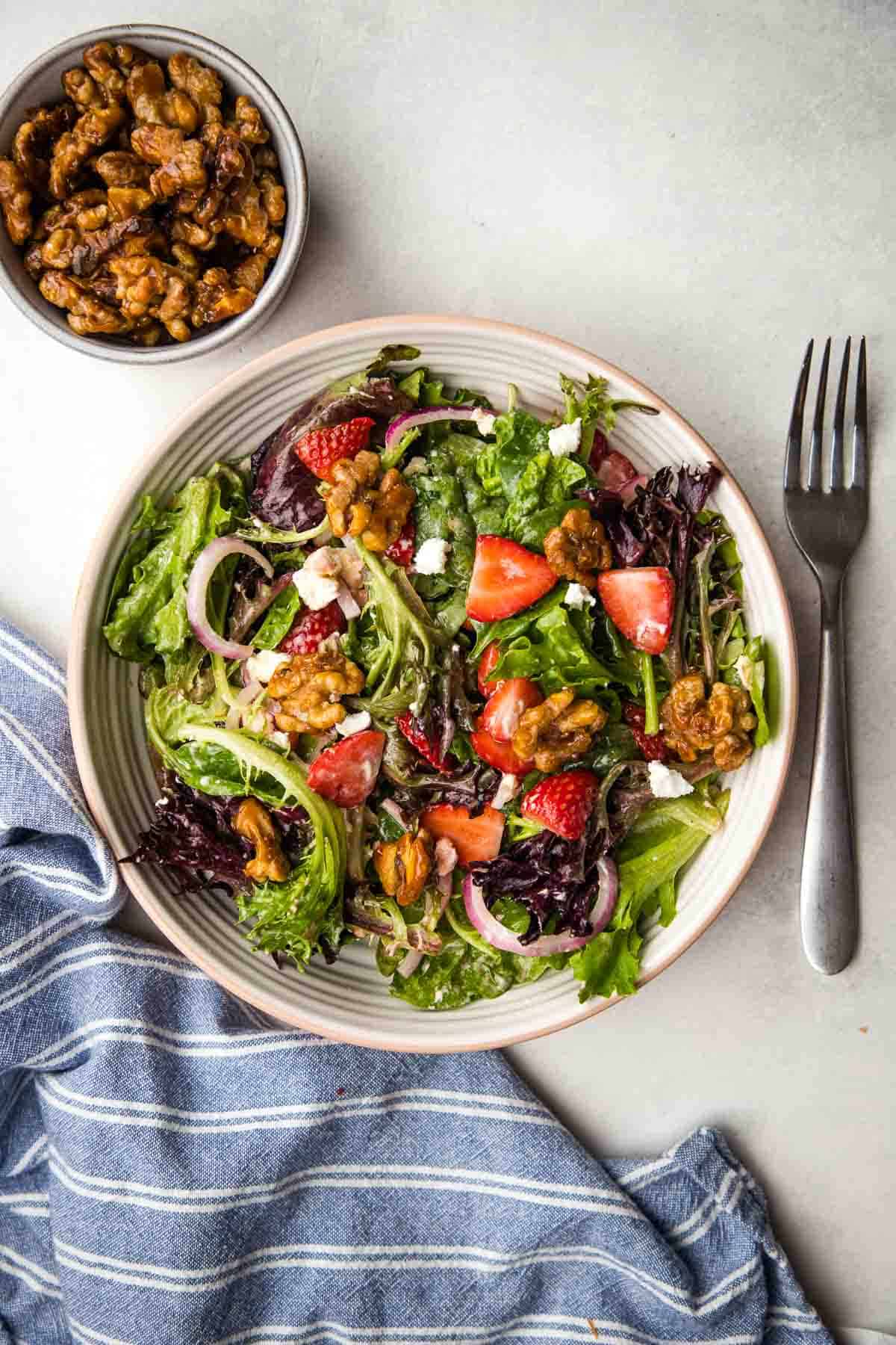 A Strawberry and goat cheese salad in a bowl with a bowl of candied walnuts on the side.