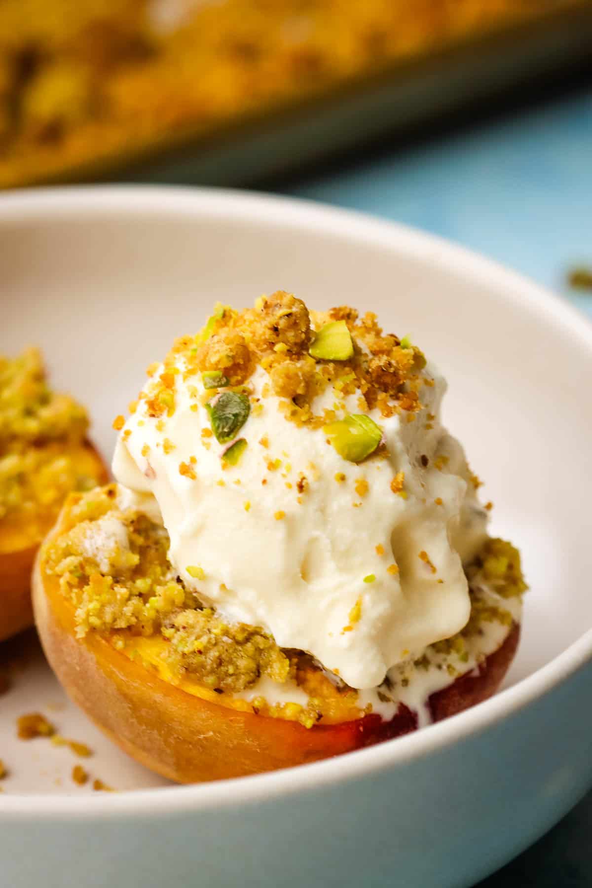 Roasted, halved peaches in a bowl with a scoop of vanilla ice cream and pistachio crumble.