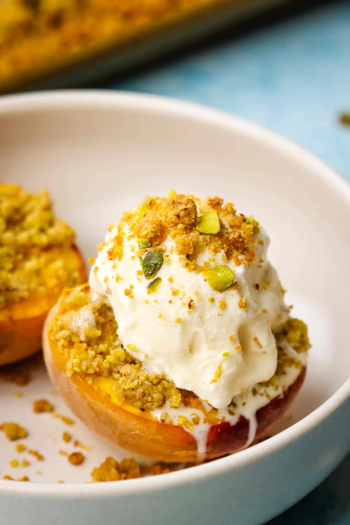 Roasted, halved peaches in a bowl with a scoop of vanilla ice cream and pistachio crumble.