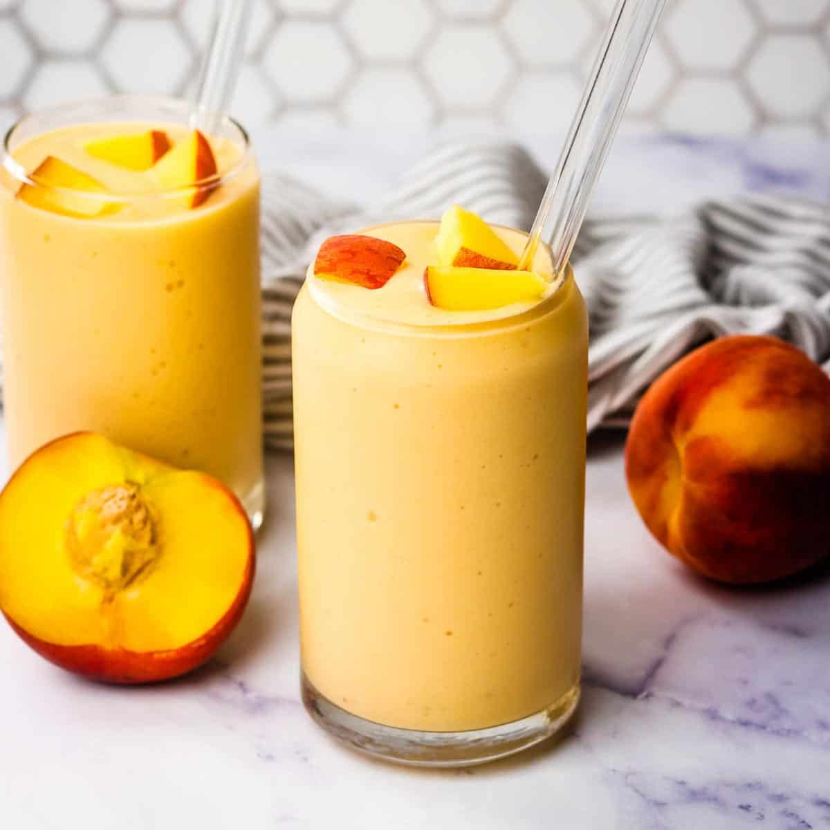Two banana peach smoothies garnished with chopped peaches and glass straws.