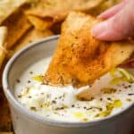 A homemade pita chip being dipped into a yogurt feta dip in a small bowl.