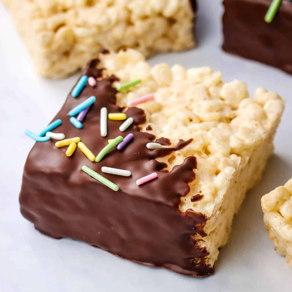 A rice krispie treat cut into a square and dipped diagonally in chocolate and then sprinkled with pastel sprinkles.
