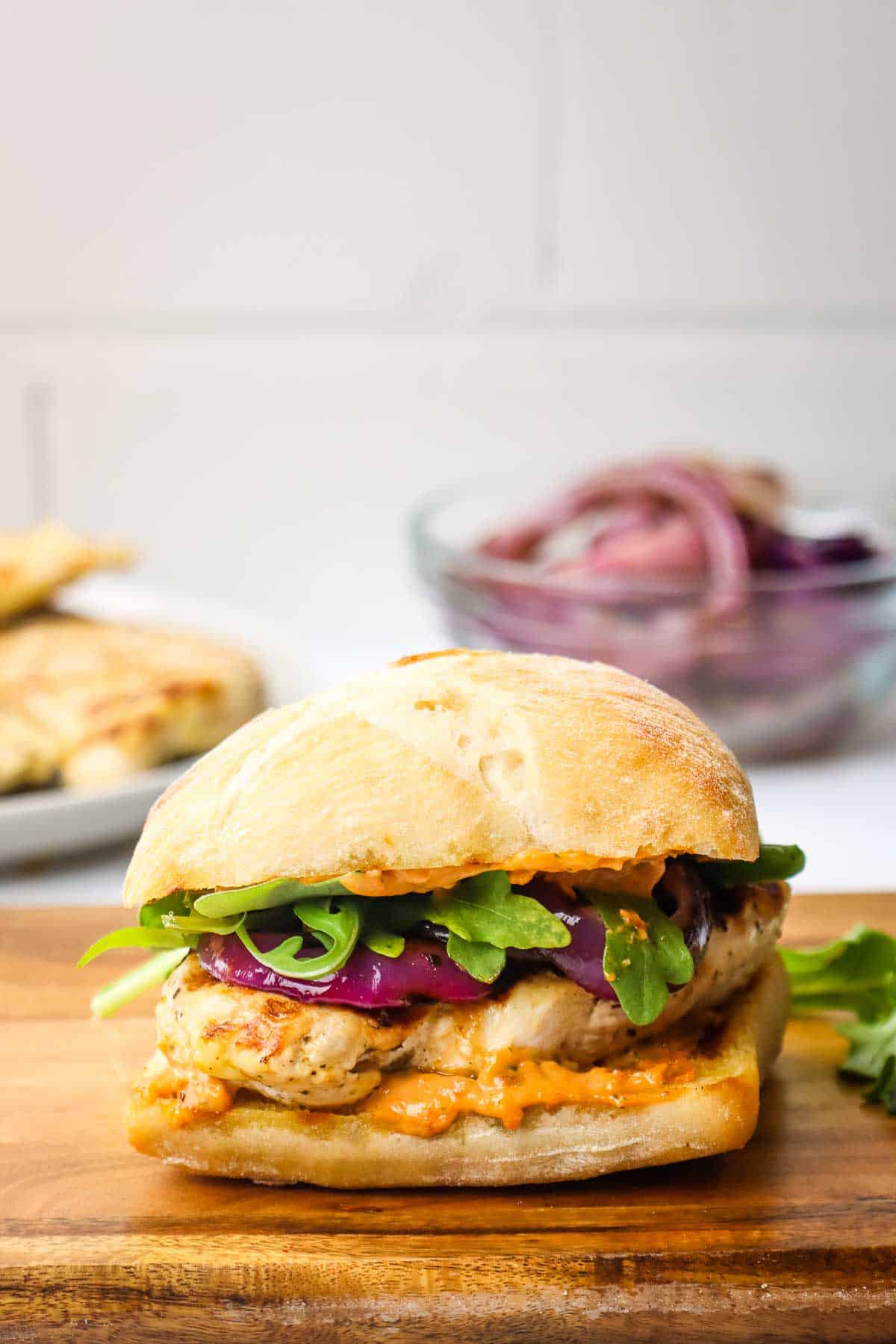 A grilled chicken sandwich in a wooden cutting board with a bowl of grilled red onion in the background.