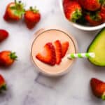 Overhead view of an avocado strawberry smoothie garnished with sliced strawberries and a green straw.