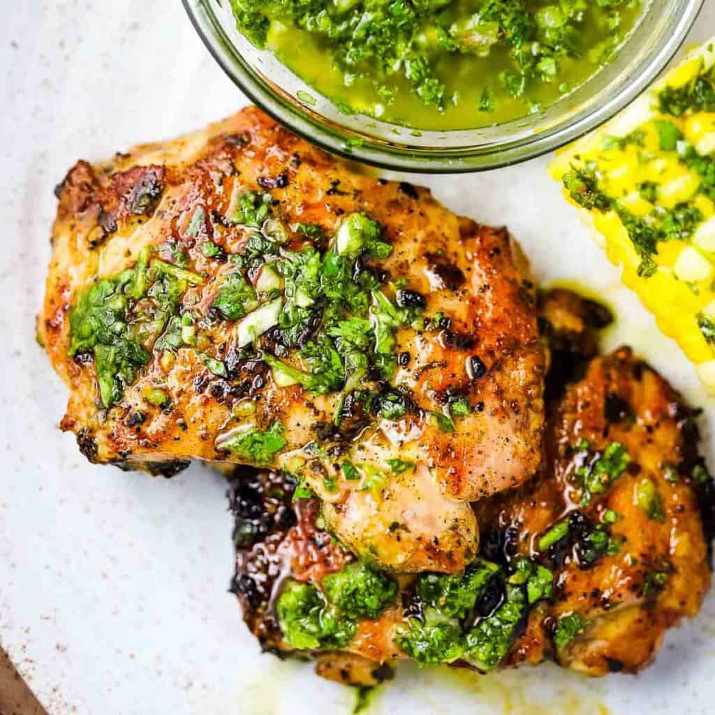 Overhead view of cooked chicken thighs slathered with chimichurri with a bowl of chimichurri in the background.