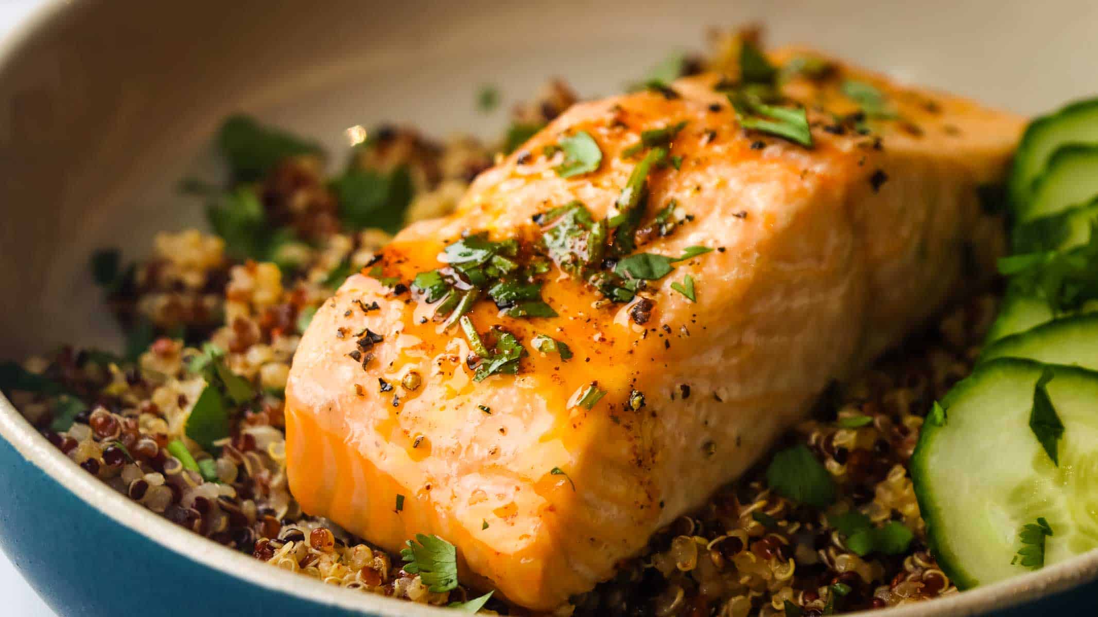 Closeup of a roasted salmon fillet on a bed of quinoa garnished with cucumber and cilantro.