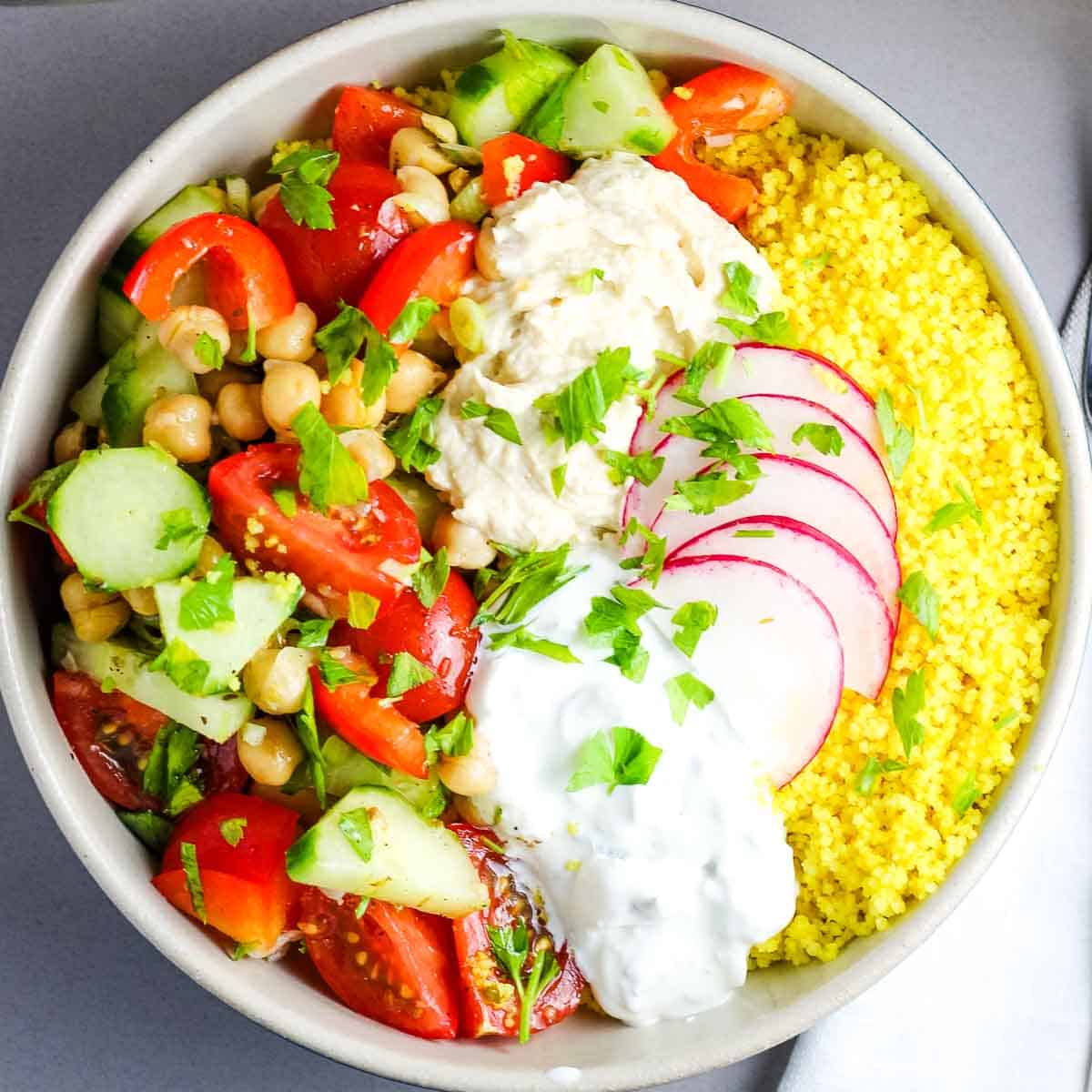 Overhead view of a bowl with couscous, chickpeas, vegetables, hummus and tzatziki, garnished with radish and parsley.