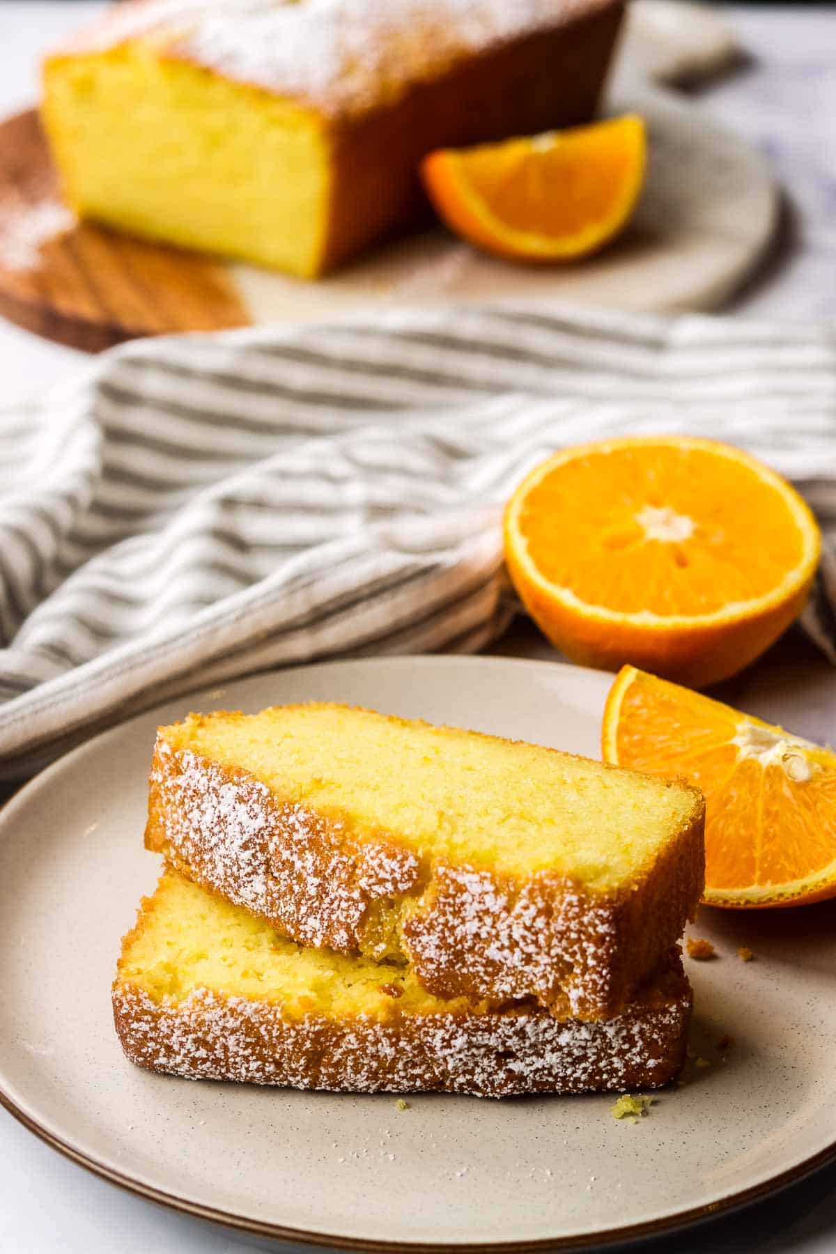 Two slices of orange loaf cake on a small place with orange wedges and halves in the background.