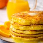 A stack of pancakes with a pat of melted butter on top and a pitcher of orange syrup in the background.