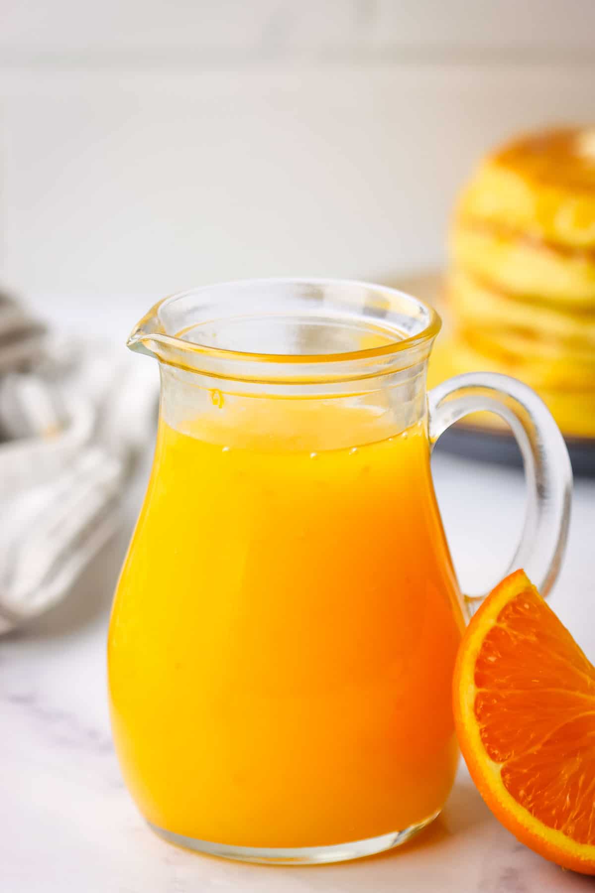 A pitcher of orange pancake syrup with a stack of pancakes in the background.