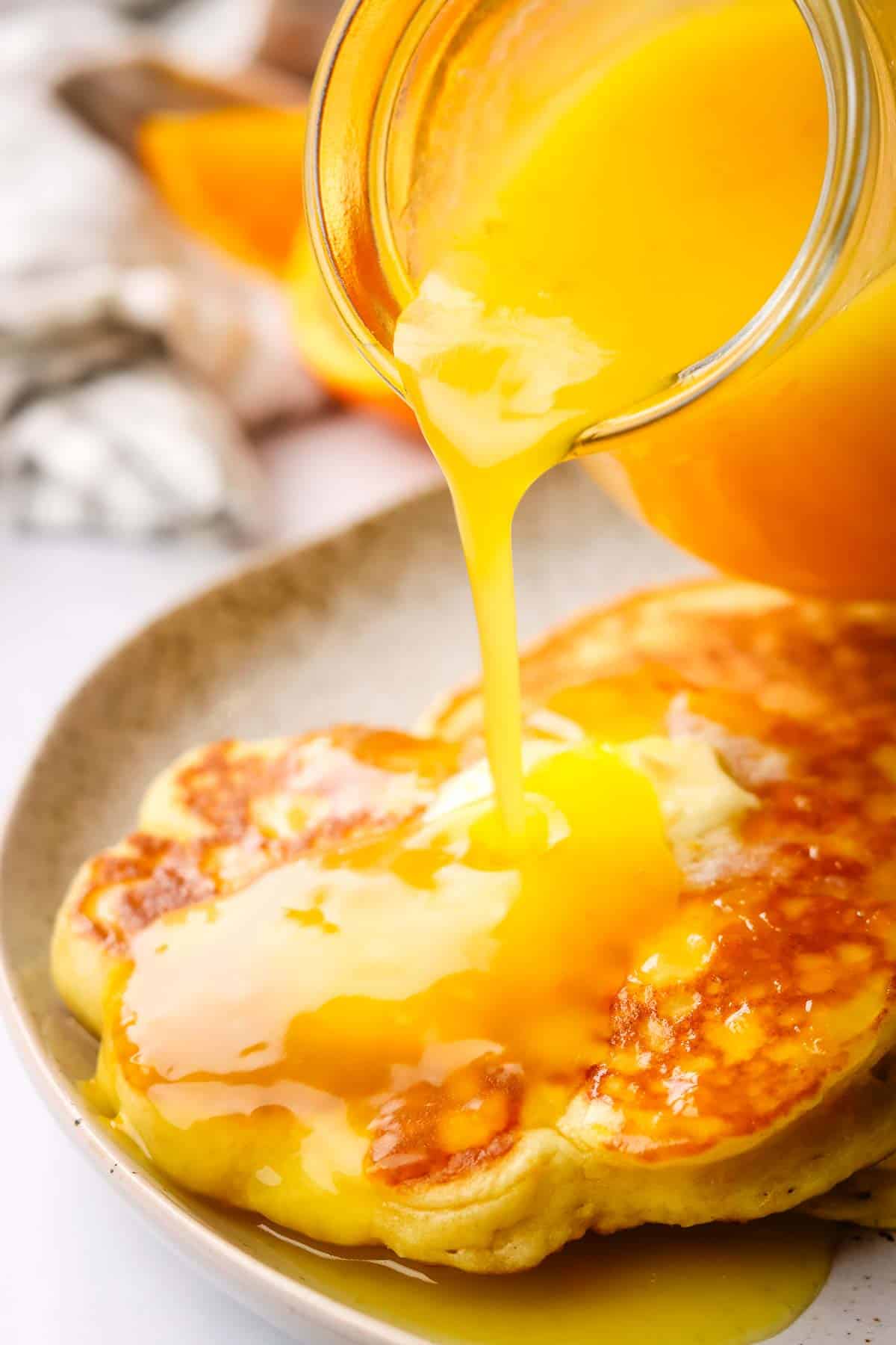 pouring orange syrup from a small jug over pancakes in a plate.
