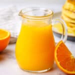 A small pitcher of orange pancake syrup with a stack of pancakes and orange slices in the background.