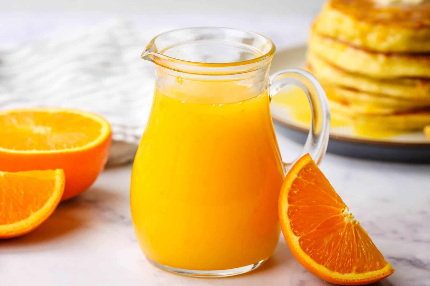 A pitcher of orange syrup with orange slices and a stack of pancakes in the background.
