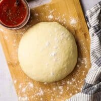 A ball of raw pizza dough on a wooden cutting board with a jar of pizza sauce in the background.