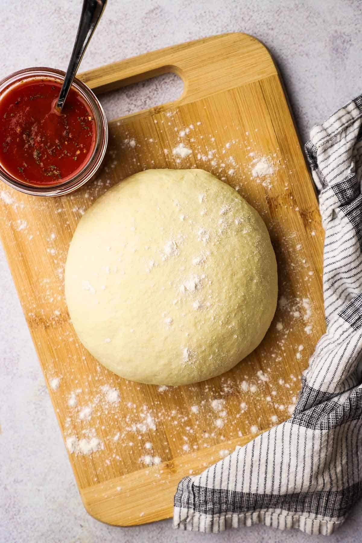 A ball of raw pizza dough on a wooden cutting board with a jar of pizza sauce in the background.