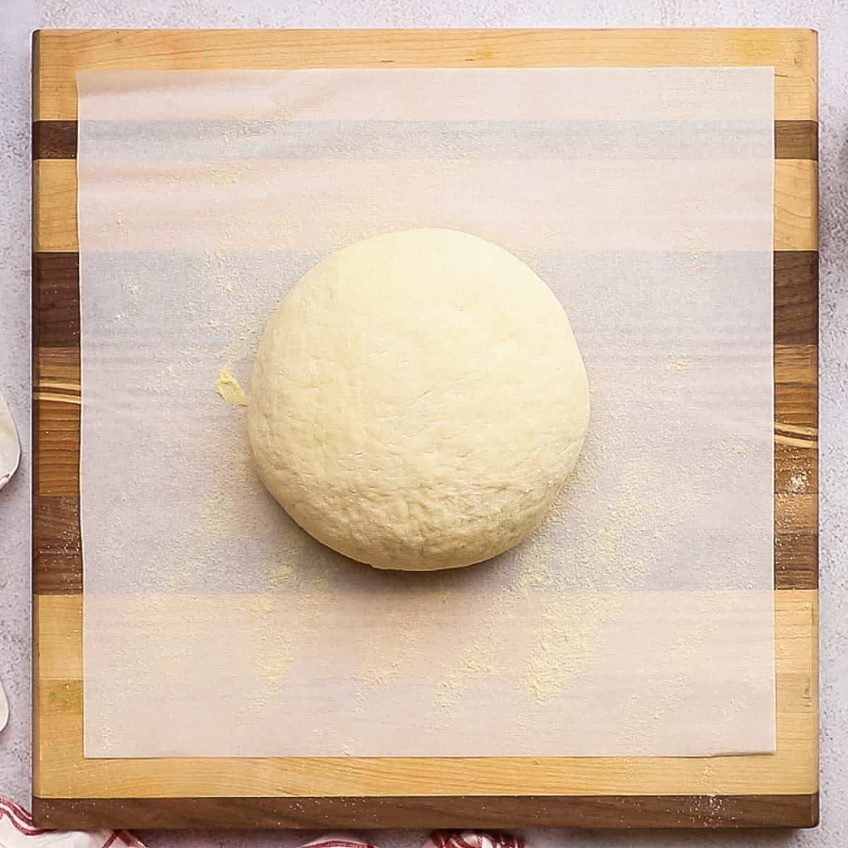 A ball of ready pizza dough on a parchment lined wooden cutting board.