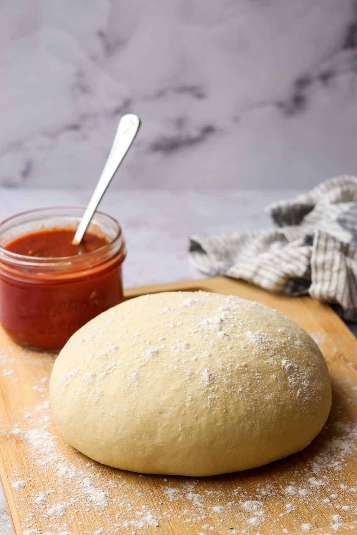 Side view of a ball of raw pizza dough sprinkled with flour on a wooden cutting board with a jar of pizza sauce in the background.