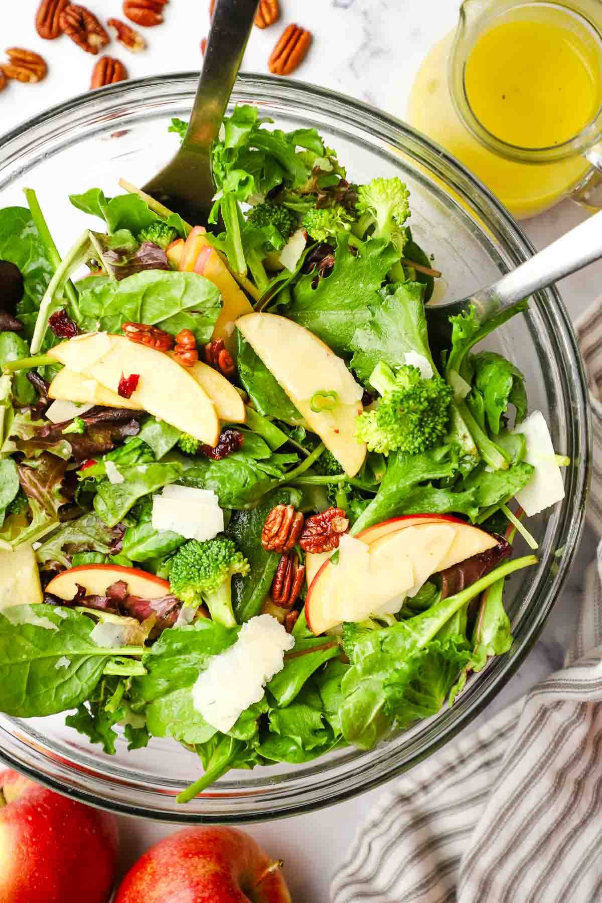A mixing bowl full of tossed salad with serving utensils. A jar of vinaigrette and apples in the background.