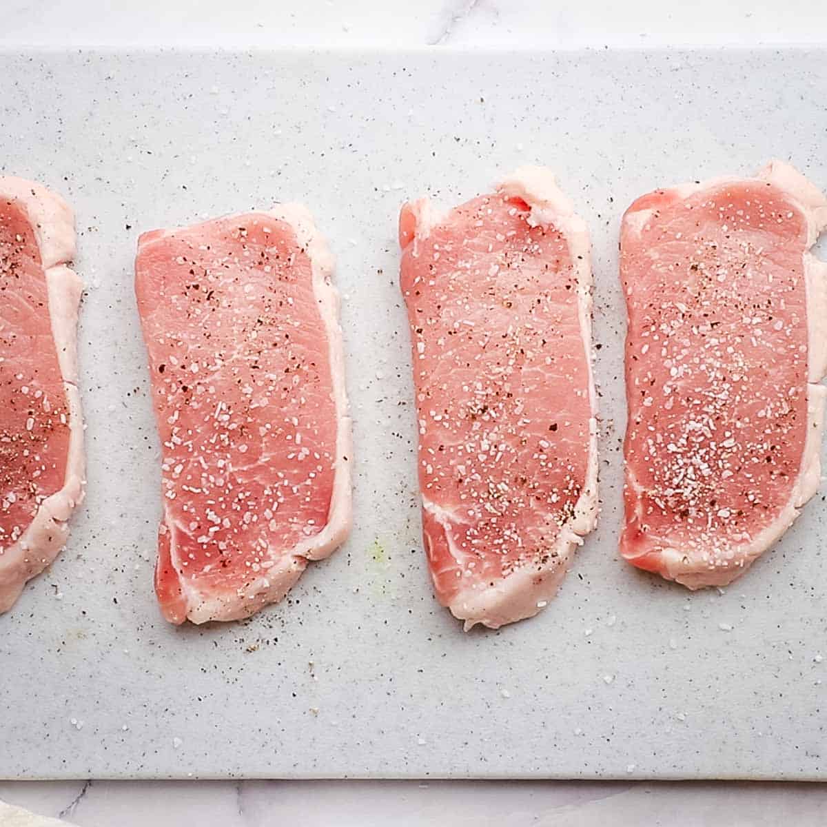 four pork chops on a cutting board with salt and pepper on them