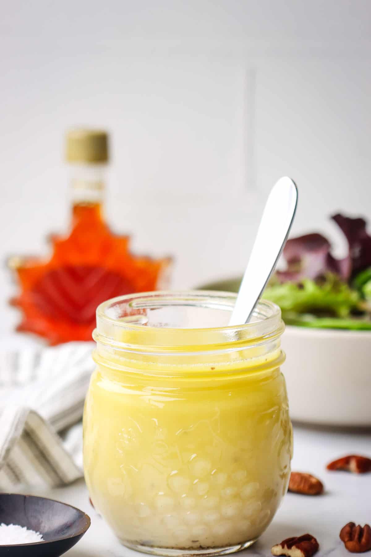 A jar of homemade vinaigrette with a spoon in it and a salad and maple syrup bottle in the background.