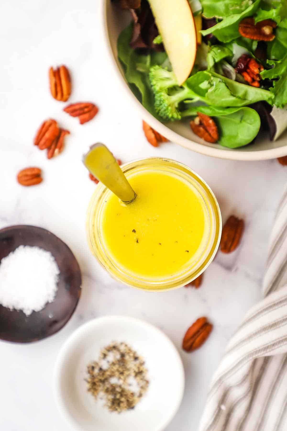 an overhead view of a jar of maple vinaigrette with a spoon in it and a bowl of salad, pecans, and few spices in the background