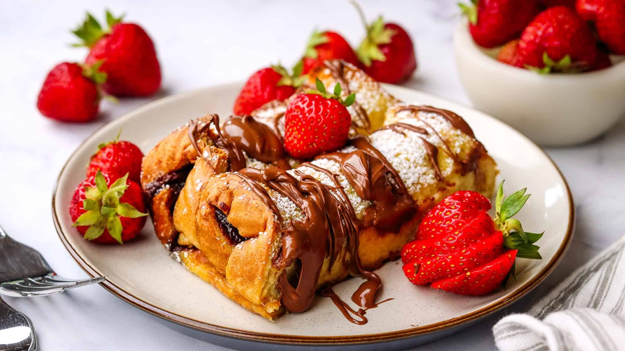 A plate of rolled nutella French toast garnished with fresh strawberries, with one roll sliced open to show the inside.