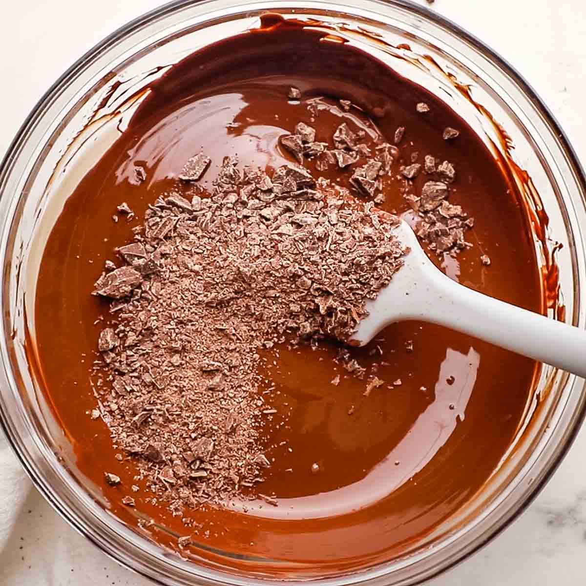 A bowl of melted chocolate sprinkled with unmelted chocolate and a rubber spatula for stirring.