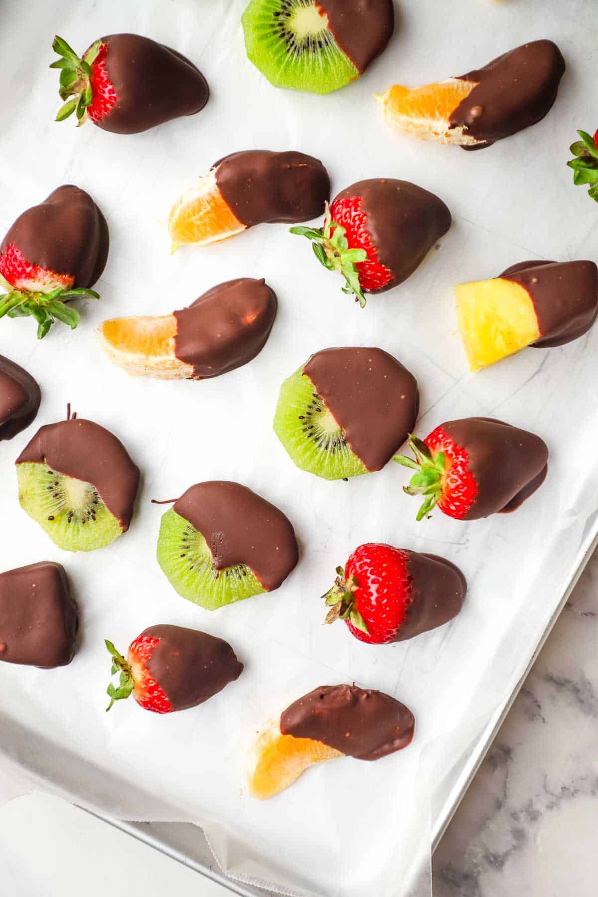 Fruits dipped in chocolate and drying on a parchment lined baking sheet.