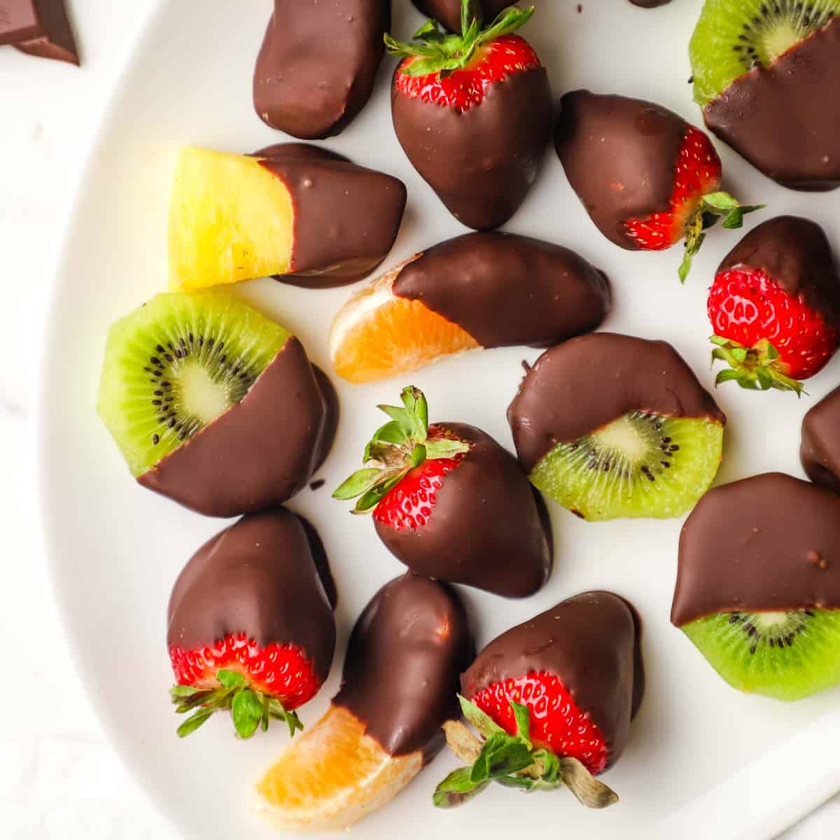 Close up view of a platter of various fruits dipped in dark chocolate on a white oval platter.