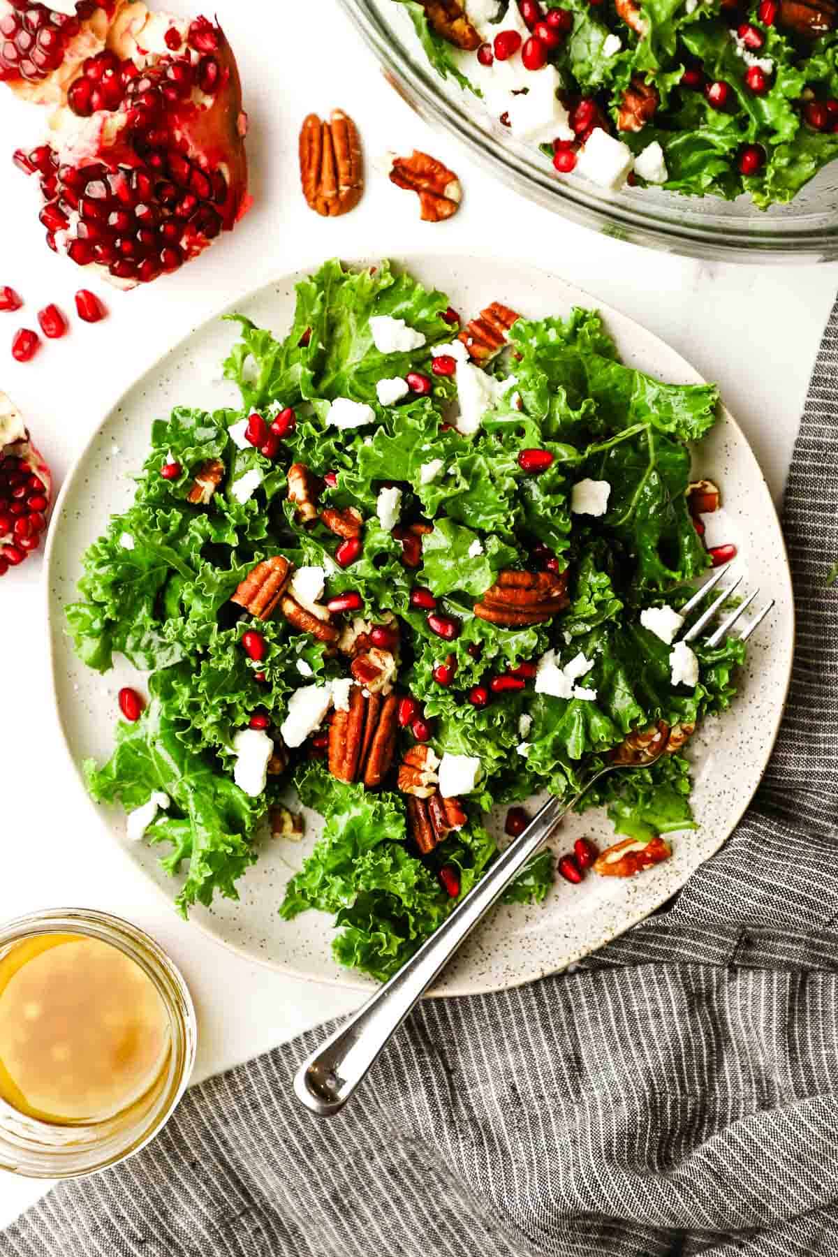 Overhead view of a plate of kale salad topped with pomegranate arils and feta cheese and pecans.