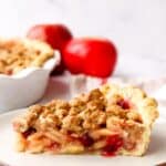 Side view of a slice of apple cranberry pie with crumble topping on a plate with red apples in the background.