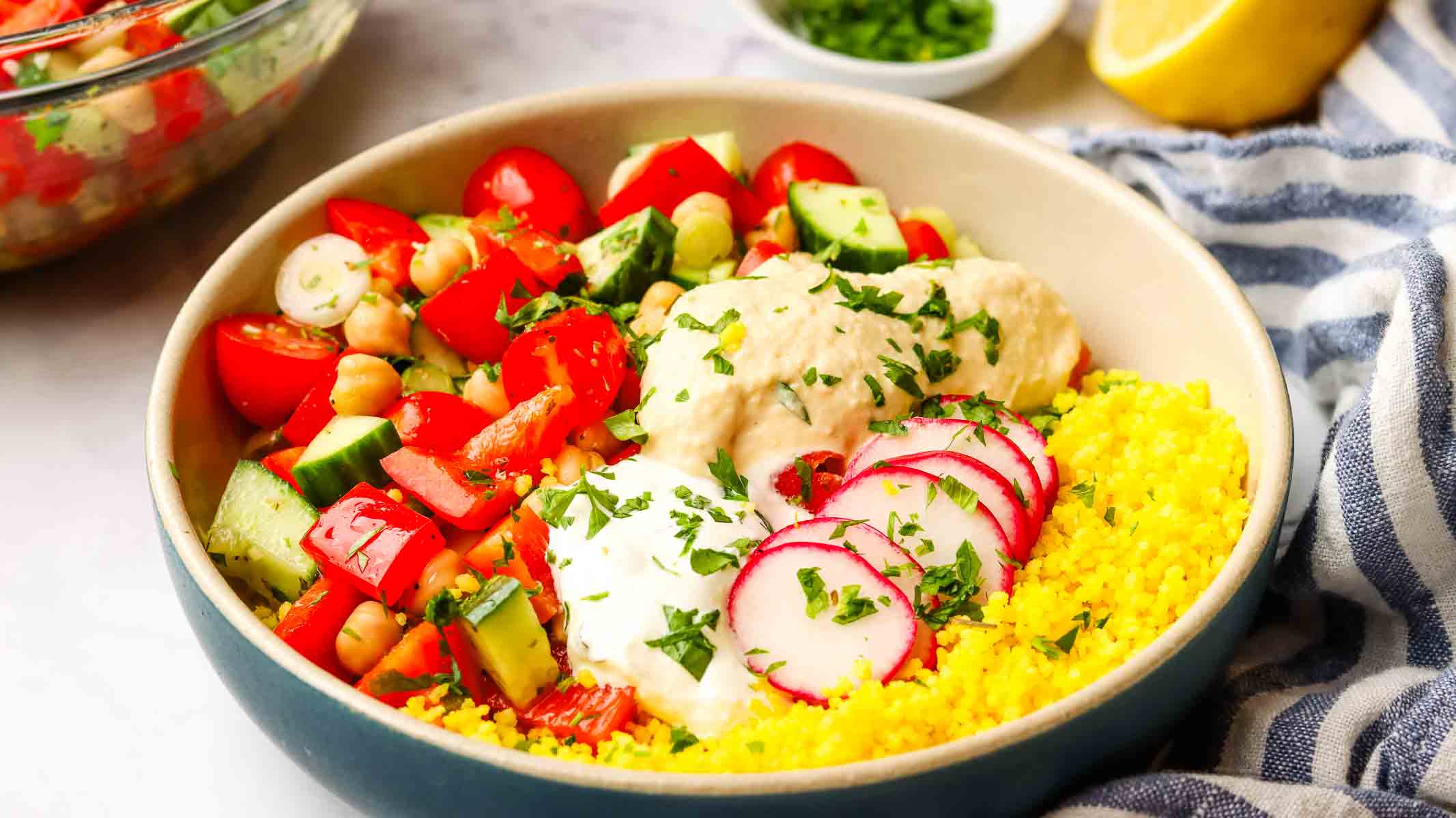 an overhead view of a colorful Mediterranean bowl with couscous, fresh vegetables, chickpeas, hummus, and tzatziki