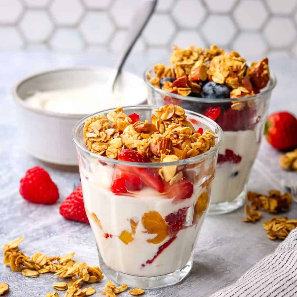 Two breakfast parfaits in glasses with a bowl of yogurt in the background.