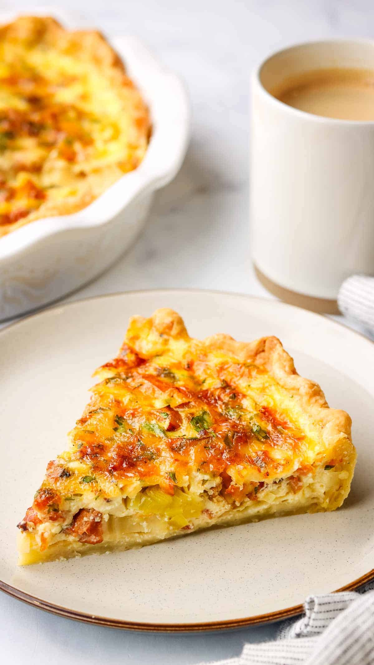 a slice of bacon and leek quiche with a dish of quiche and coffee mug in the background