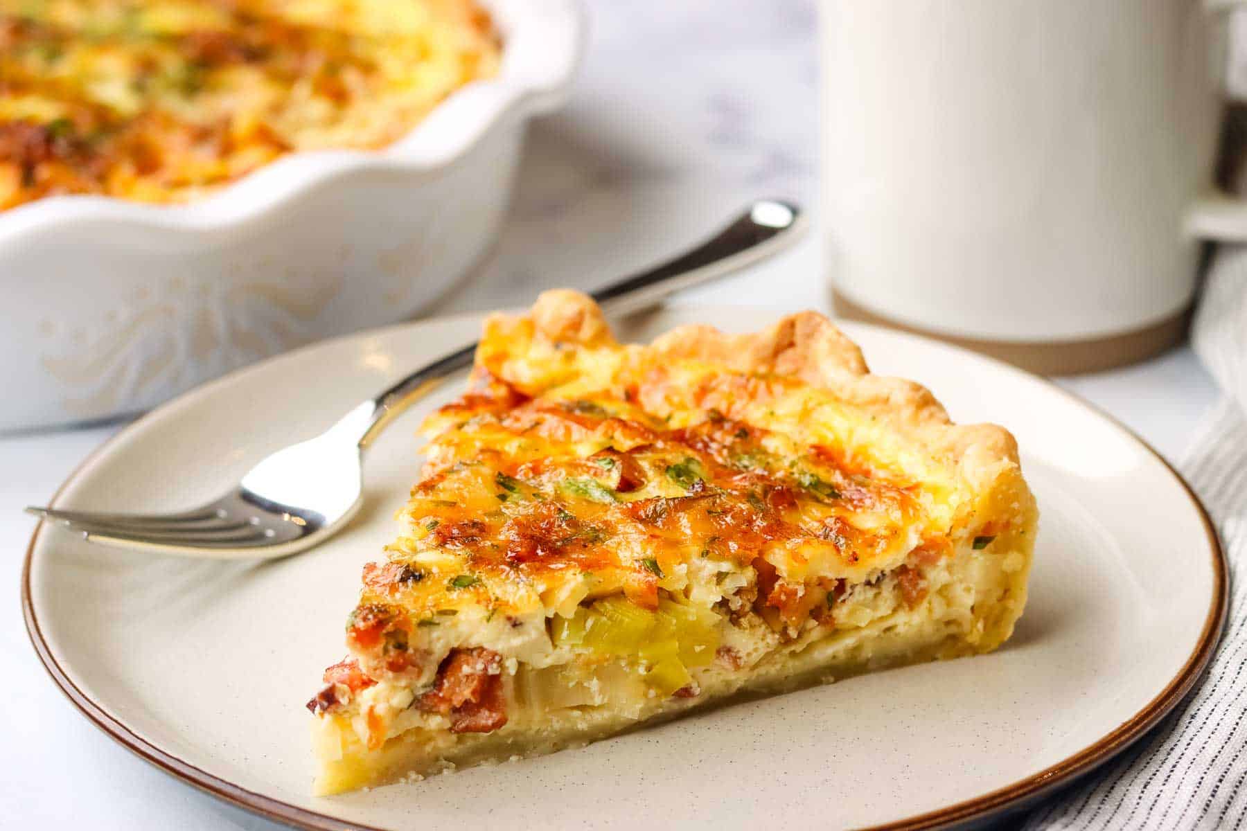 a slice of bacon and leek quiche with a dish of quiche and coffee mug in the background